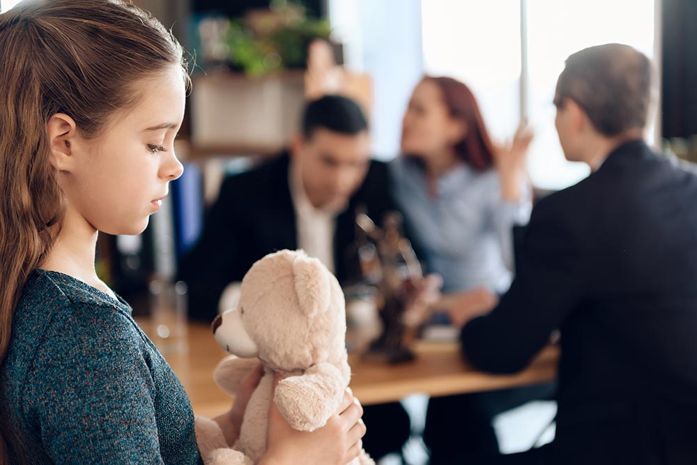 Une petite fille avec une pelluche avec en arrière plan trois adultes
